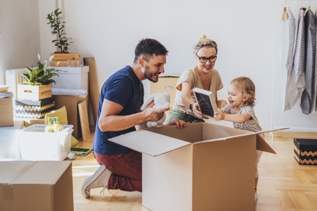 A couple and their young child unpack belongings from a cardboard box in a home, surrounded by moving boxes