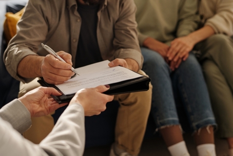A man signs a document on a clipboard while two people sit beside him during a child custody meeting. | The Law Offices of Andrea Schneider