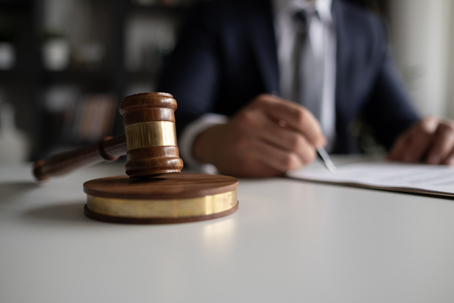 A judge’s gavel on a desk with a person in a suit signing legal documents in the background. | The Law Offices of Andrea Schneider