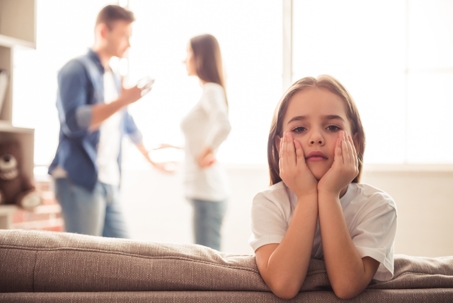 A sad young girl sits on a couch with her head in her hands while a man and woman argue in the blurred background of a home setting. | The Law Offices of Andrea Schneider